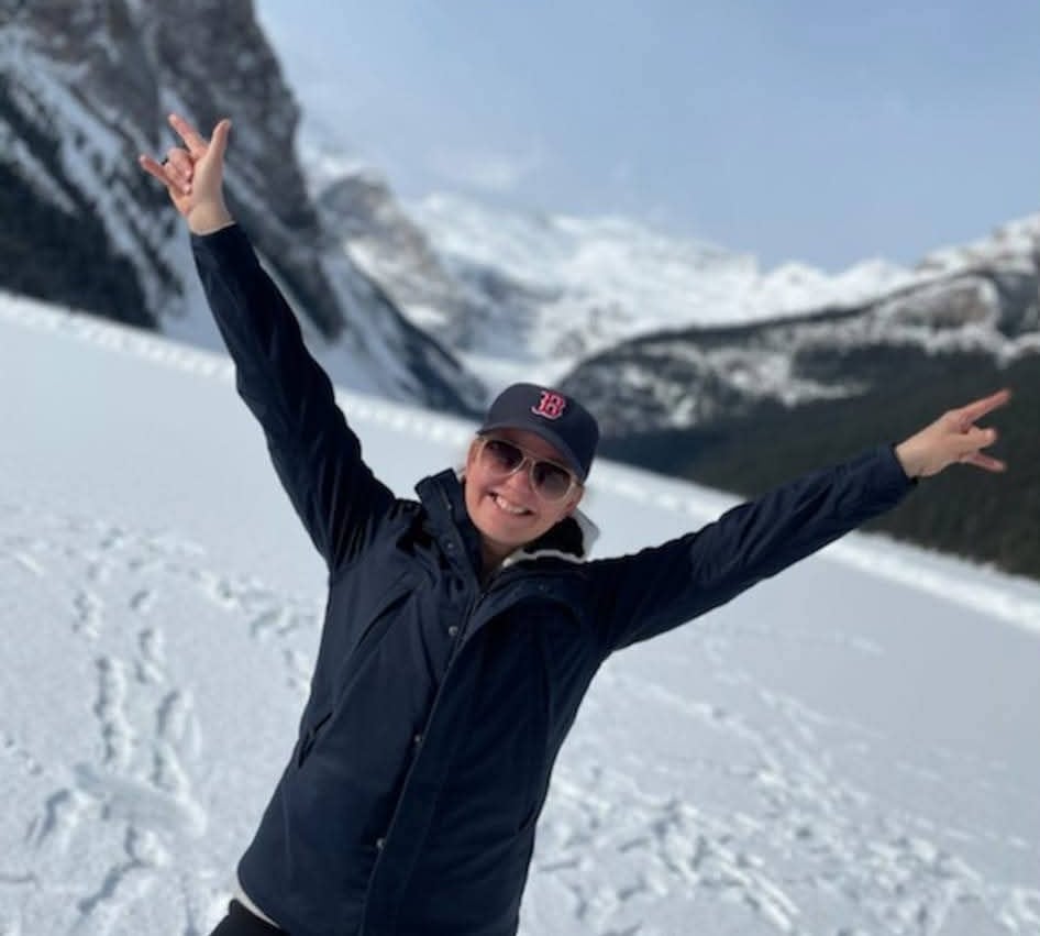 A smiling woman raises her arms joyously while standing in front of a frozen lake and snowy mountains.