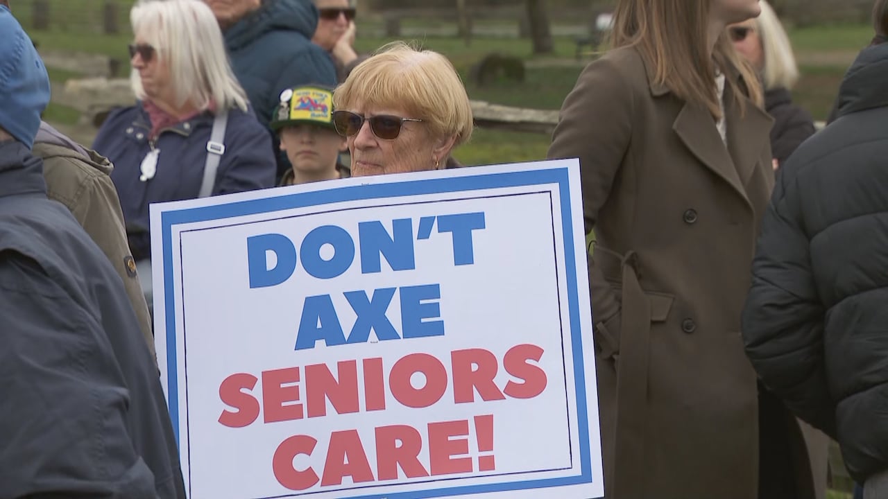 An older woman wearing sunglasses holds a sign that reads “Don’t axe seniors care” while standing among a group of people outdoors.