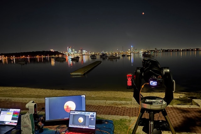 Computers and cameras gathered on the ground under a dark sky with a blood moon, with the Perth skyline below.