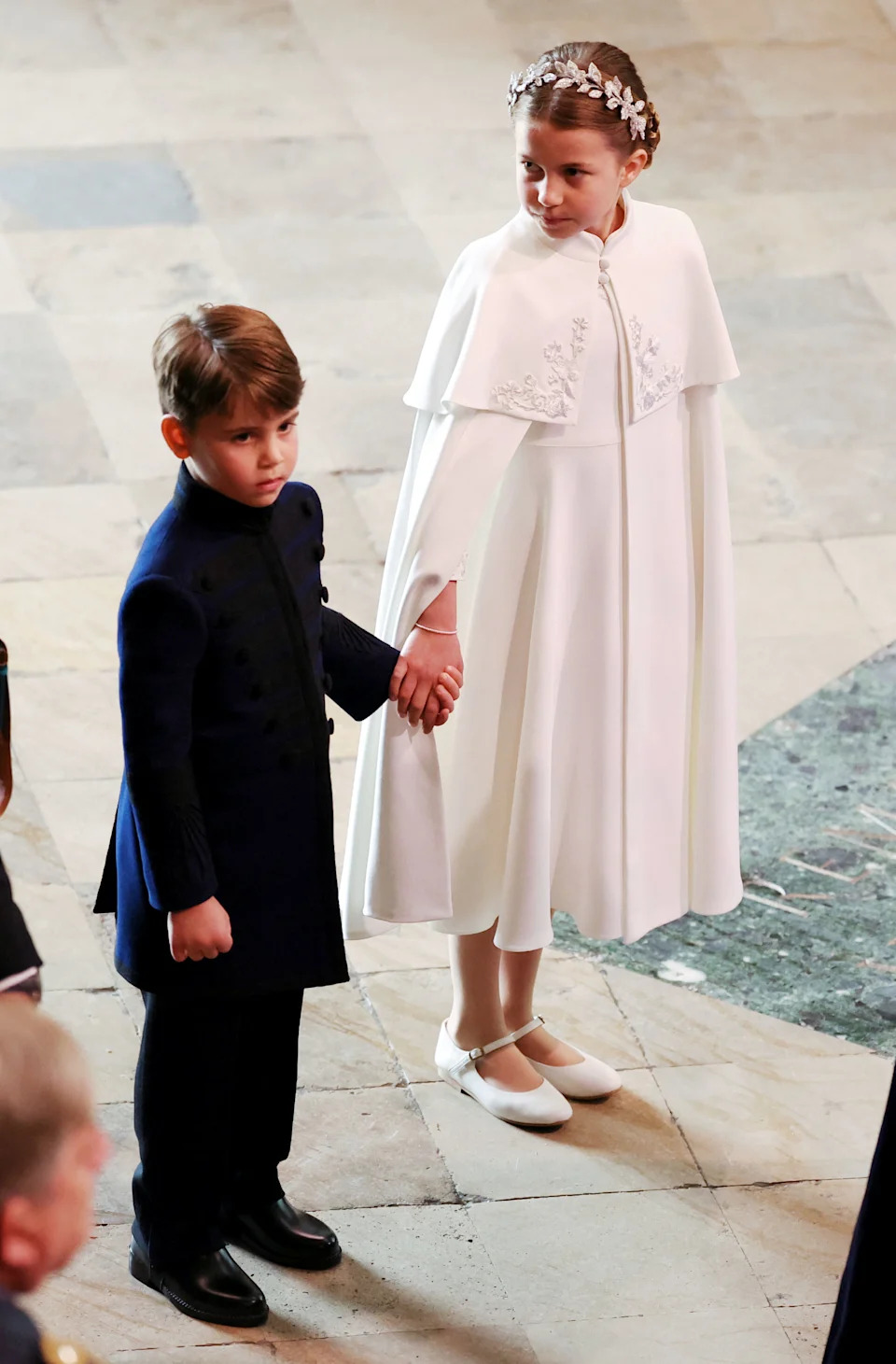 Princess Charlotte wearing a white cape holding hands with Prince Louis at King Charles's coronation