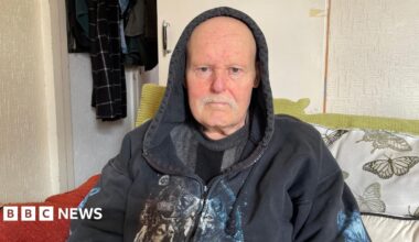 Tunde Olawoye stood behind the counter at his pharmacy wearing a brown fleece jumper. There are shelves of drugs and medicine behind him.