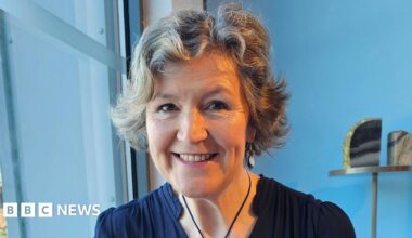 A woman in a navy blue dress smiling at the camera while sitting in front of a blue wall.