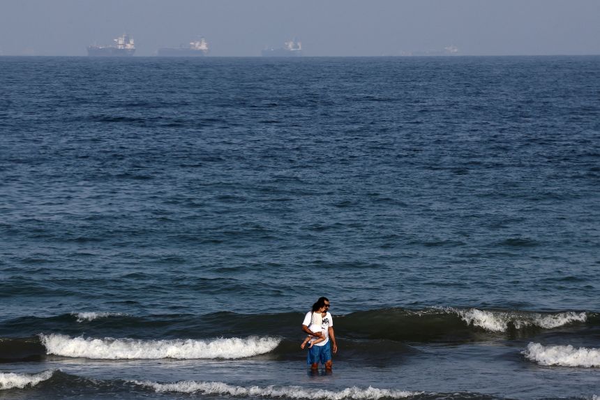 People stand in the water, with tankers in the background, as Iran vows to close the Strait of Hormuz, amid the US-Israel conflict with Iran, in Fujairah, United Arab Emirates.