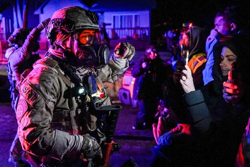 A US law enforcement officer in protective clothing and a gas mask standing in front of a hooded protester with a phone.