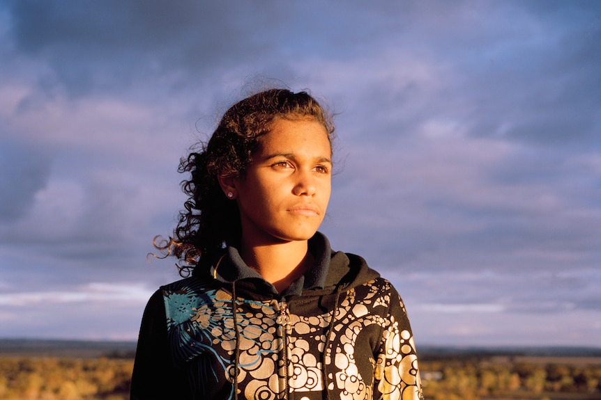 Young woman in hoodie stands outside in a scrubby landscape, with dark clouds behind her