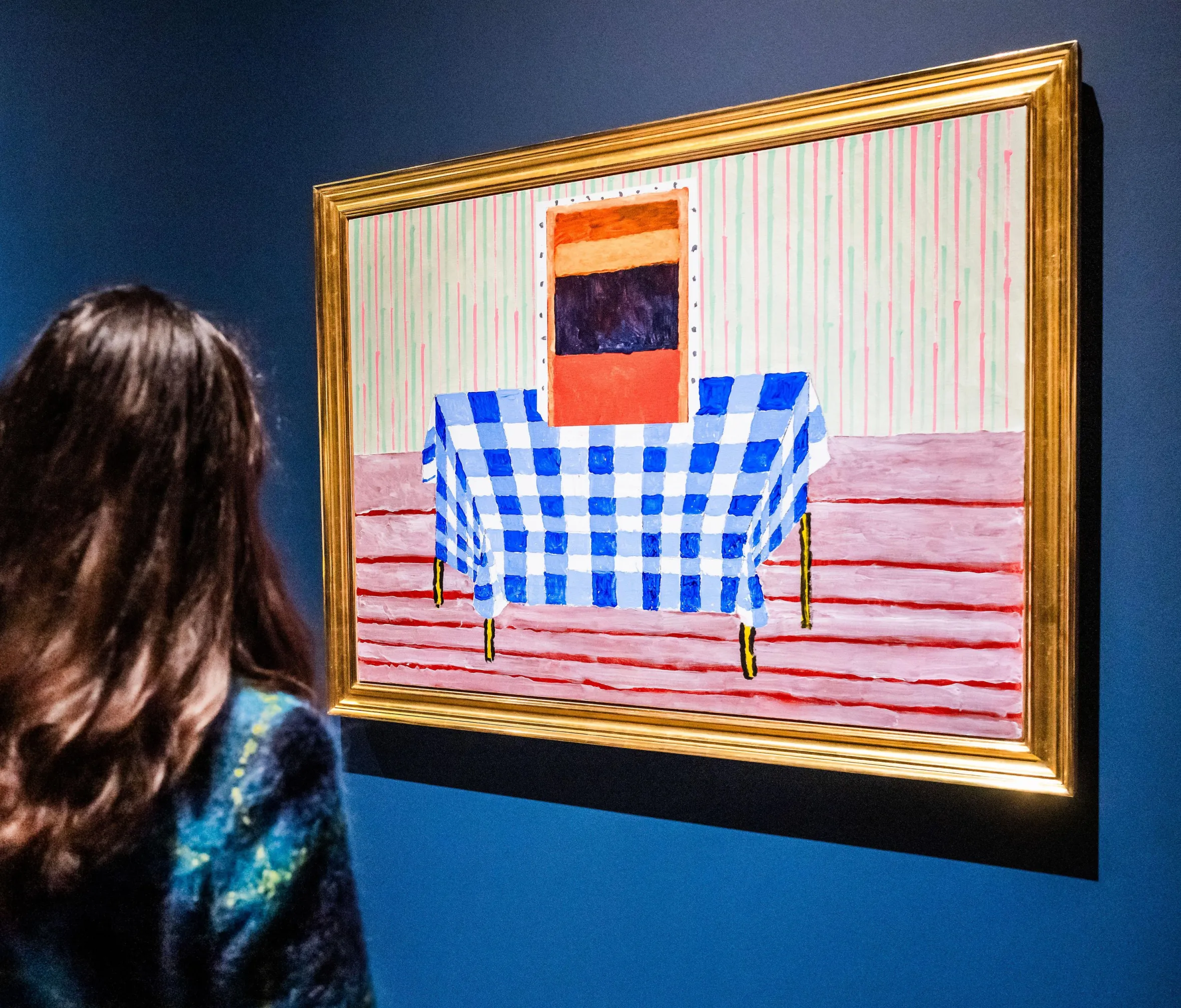 Two paintings featuring checkered tablecloths hang on a dark blue wall, viewed by a person with long brown hair.