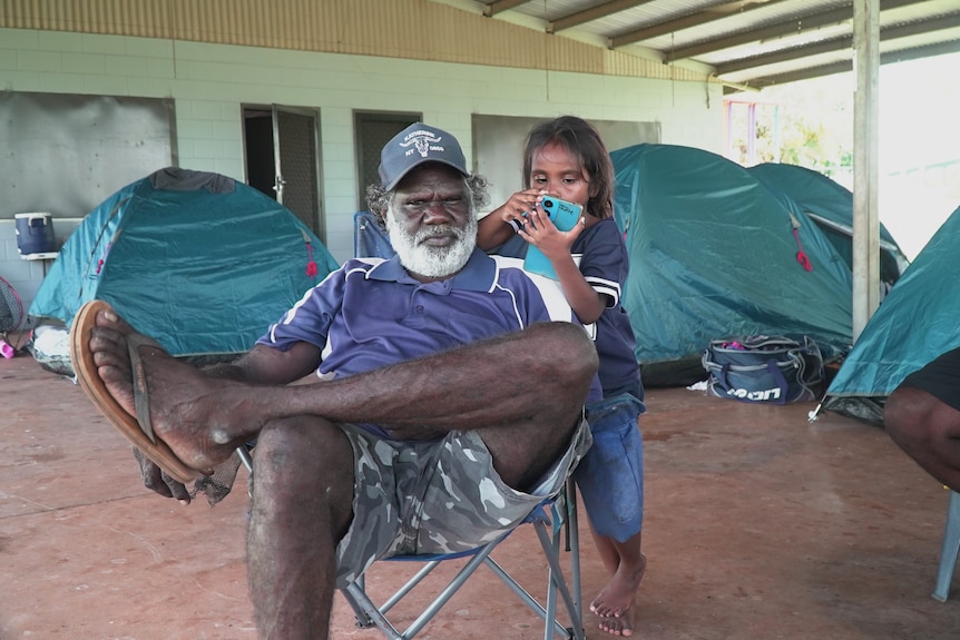 An indigenous man sitting in a camp chair with young girl standing behind 