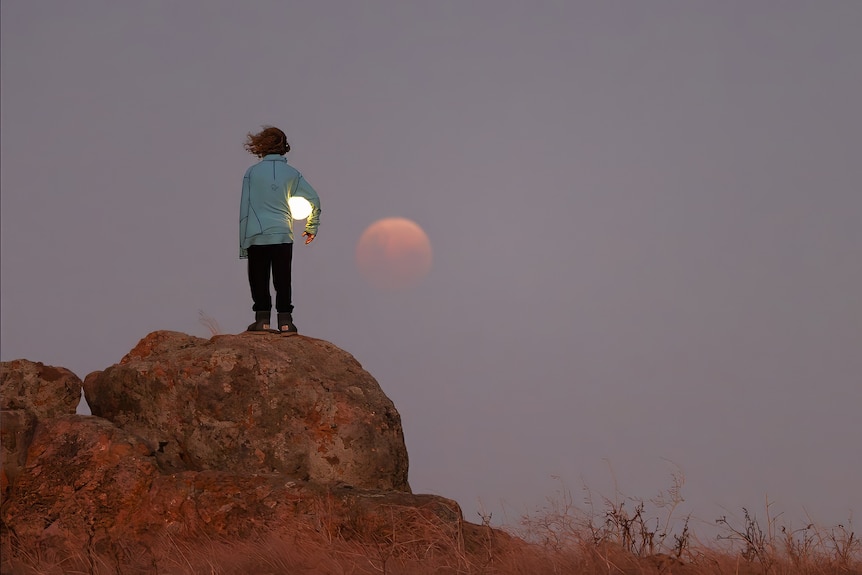 A boy stands on a large rock looking at a large blood moon in the sky.