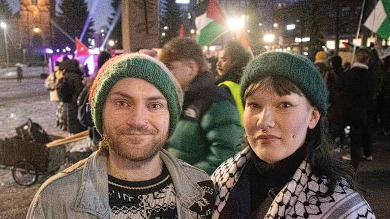A young couple wearing knit caps stand in an urban square on a winter night while others hold Palestinian flags behind them.