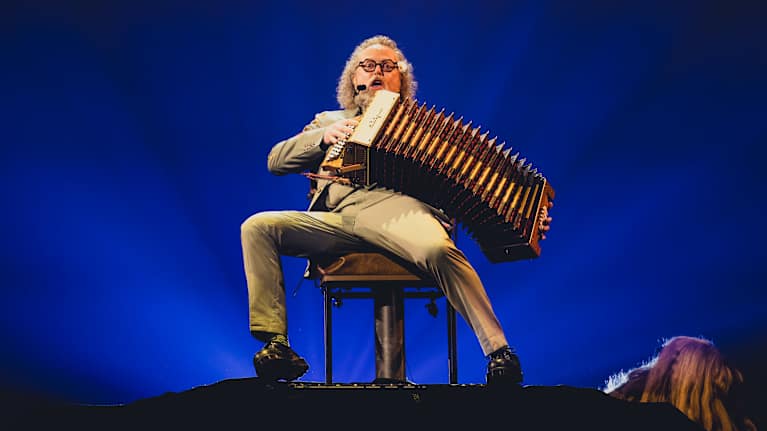 A middle-aged man wearing glasses and a beige suit sits playing an accordion onstage against a blue background.