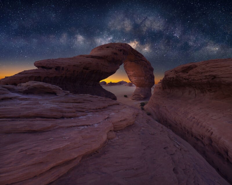 A natural sandstone arch stands under a starry night sky with the Milky Way visible above, surrounded by rocky formations and a faint orange glow on the horizon.