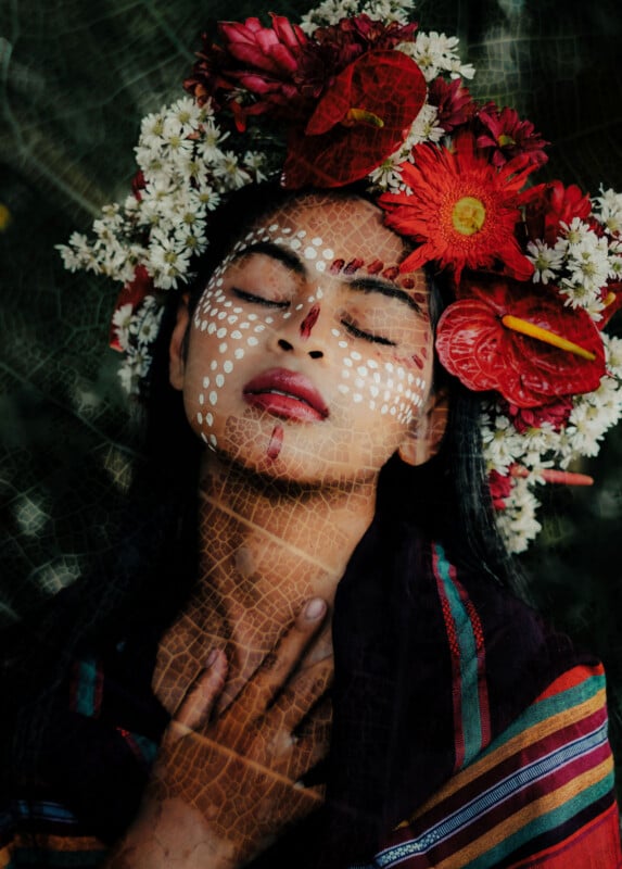 A person with eyes closed wears a crown of red and white flowers and has white face paint in dotted patterns. They touch their neck gently, dressed in colorful striped fabric. Light filters through, creating a textured overlay.