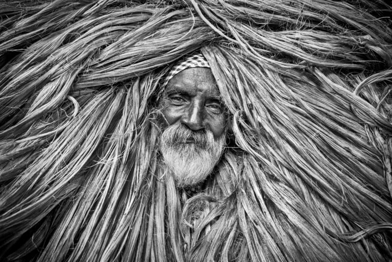 Black and white photo of an elderly man with a beard, partially covered and surrounded by long, thick strands of dried grass or fibers, creating a striking frame around his face.