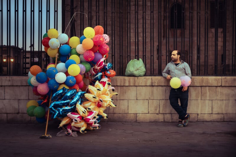 A man leans against a stone wall beside a fence, holding a pink balloon. Next to him is a large, colorful bunch of balloons and inflatable toys. A green bag rests on the wall behind him.