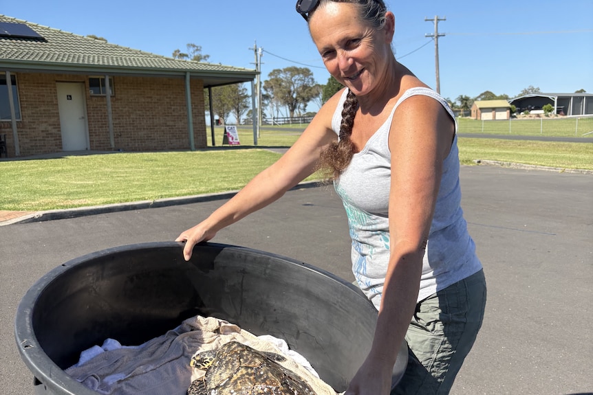 A woman in a singlet top carries a large tub with a turtle inside it