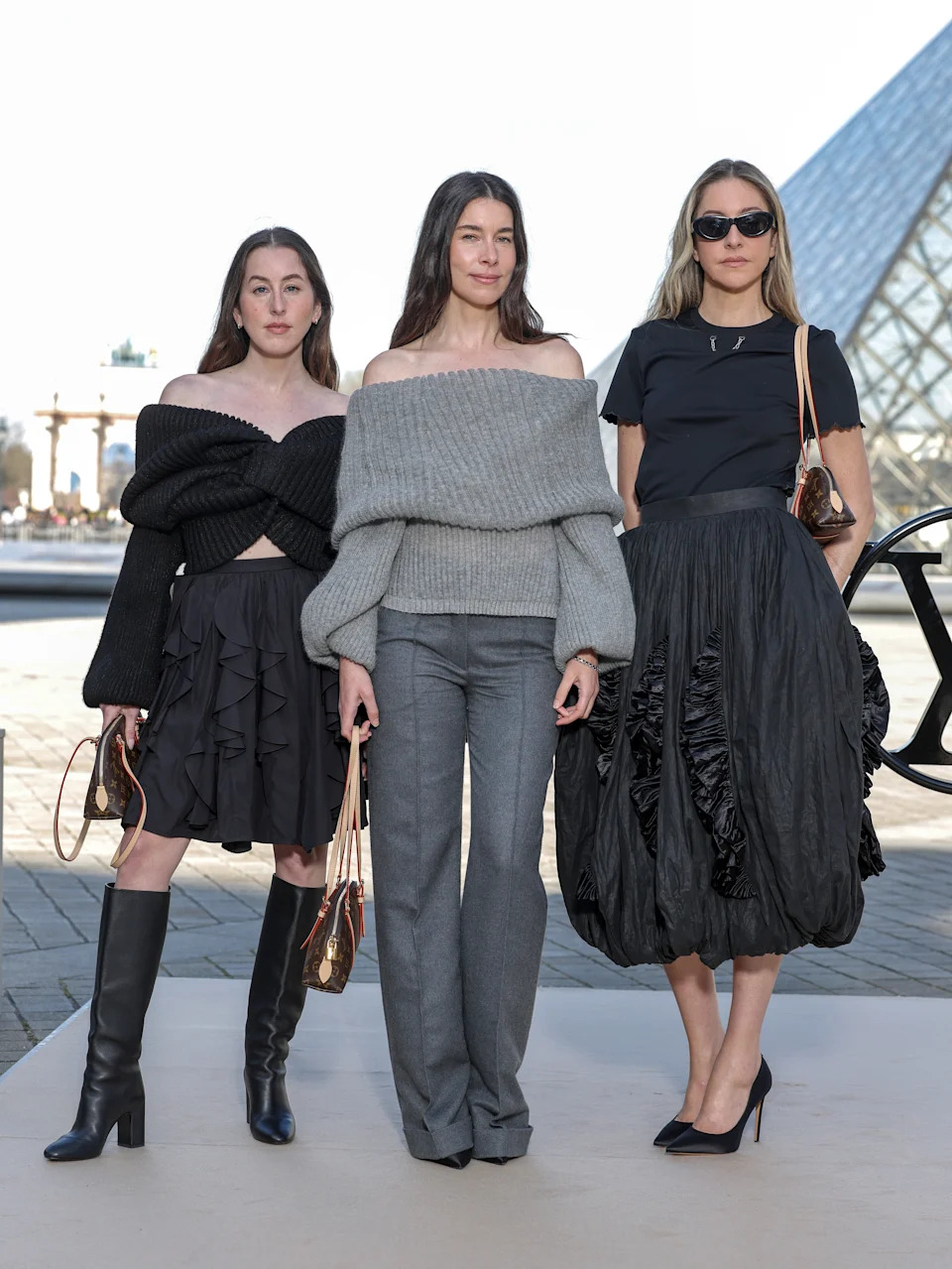 Three women in chic outfits stand together; one wears a dark off-shoulder top and skirt, another in a grey sweater and pants, the third in a black top and skirt