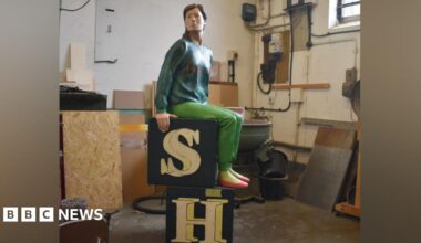 A restored sculpture shows a seated human figure positioned on two stacked black blocks with large cream‑coloured letters “S” and “H.” The sculpture is placed inside a workshop with tools, cables, wooden panels and machinery visible in the background. The figure wears a long‑sleeved top, trousers and shoes.