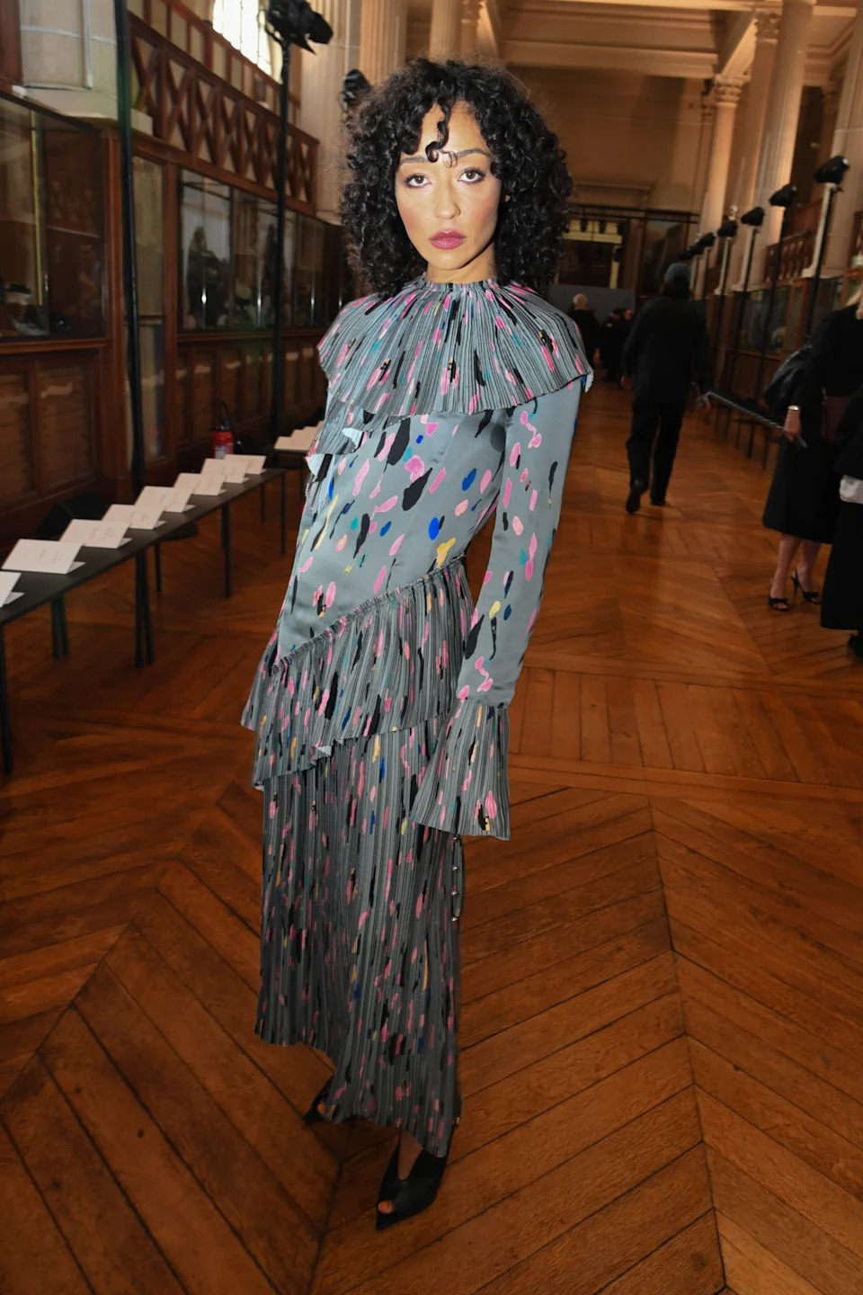 Person in a pleated, patterned dress with ruffled collar and cuffs, standing in a formal indoor venue with parquet flooring and seating