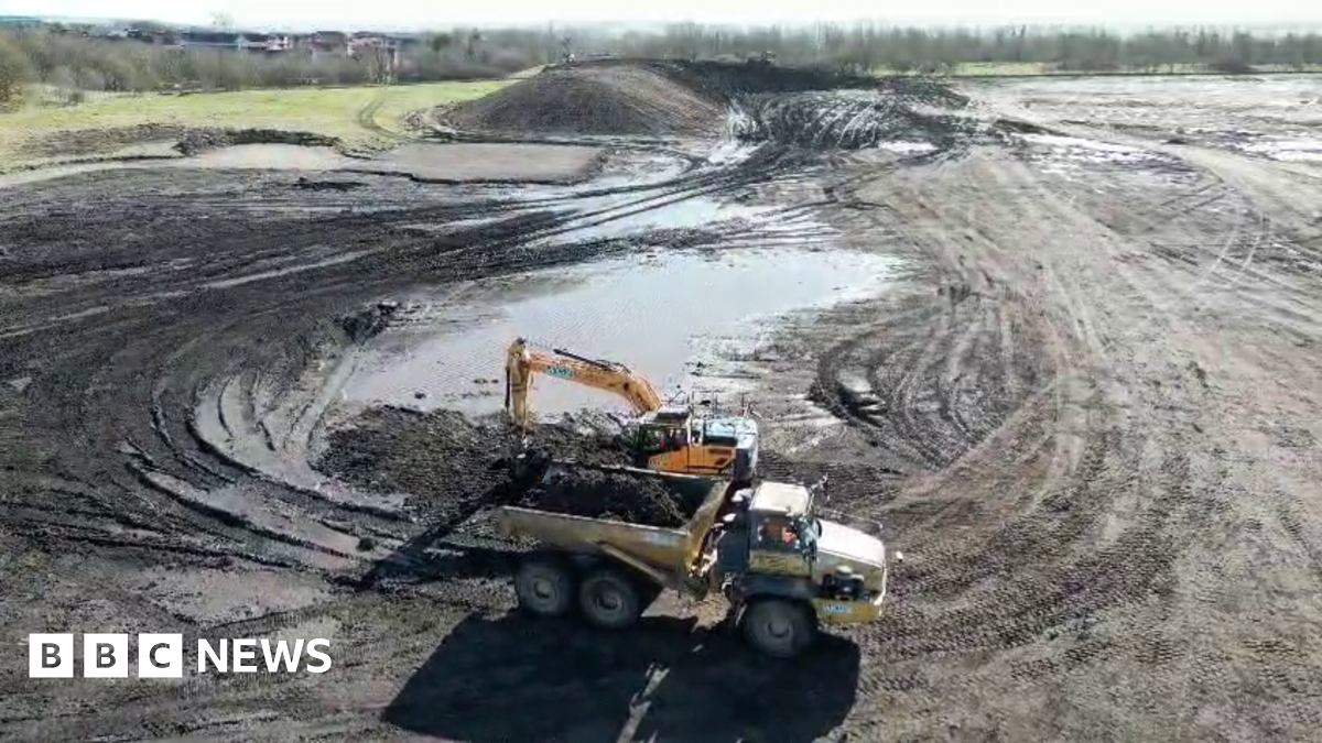 A lorry and a yellow digger at work on the muddy site of the new hospital. There are large puddles.