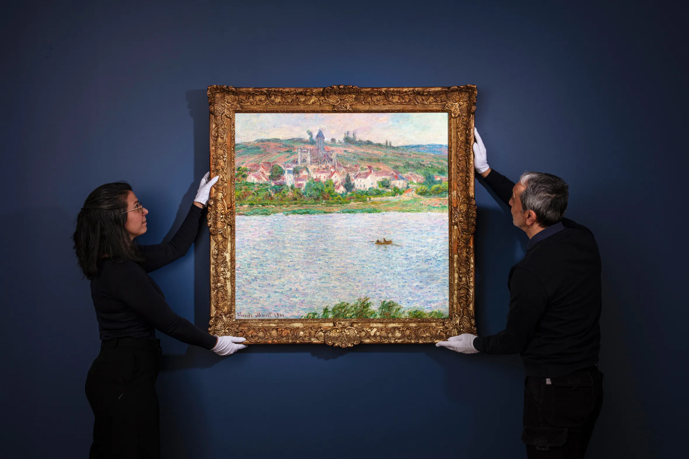 Two people in white gloves hold up a framed Claude Monet painting of a village and river.