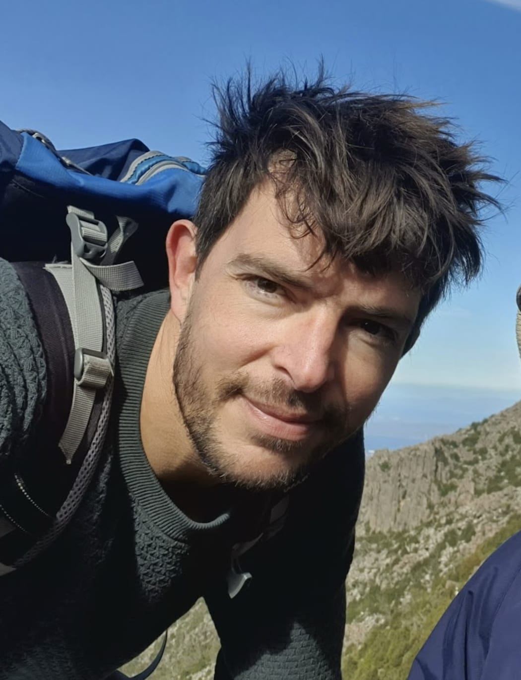 A candid selfie of marine scientist Lennart Bach, wearing hiking clothes and a backpack against a backdrop of cliffs with outcrops of bush.