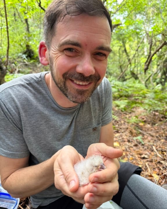 Adam is crouched down, smiling at the camera and holding a tiny white kākāpō chick in his cupped hands.