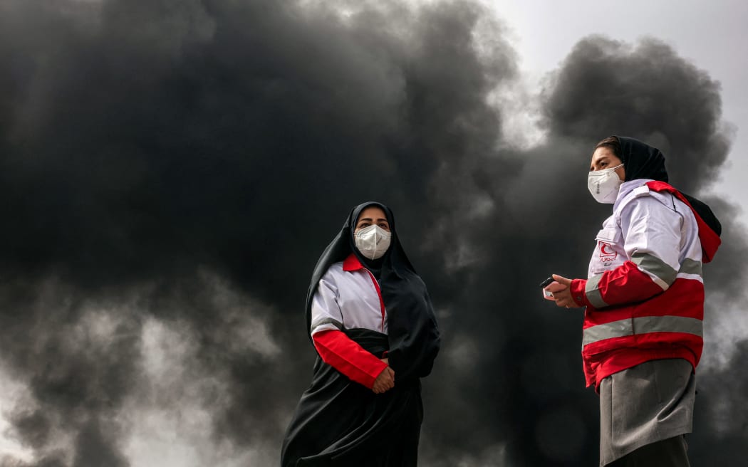 Women members of Iran's Red Crescent society stand near smoke plumes from an ongoing fire following an overnight airstrike on the Shahran oil refinery in northwestern Tehran on March 8, 2026. The United States and Israel launched strikes against Iran on February 28, sparking swift retaliation by the Islamic republic which responded with missile attacks across the region. The war has dragged in global powers, upended the world's energy and transport sectors, and brought chaos to even usually peaceful areas of the volatile region. (Photo by AFP)