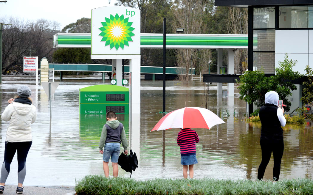 People stand next to a flooded petrol station due to torrential rain in the Camden suburb of Sydney on July 3, 2022. - Thousands of Australians were ordered to evacuate their homes in Sydney on July 3 as torrential rain battered the country's largest city and floodwaters inundated its outskirts. (Photo by Muhammad FAROOQ / AFP)