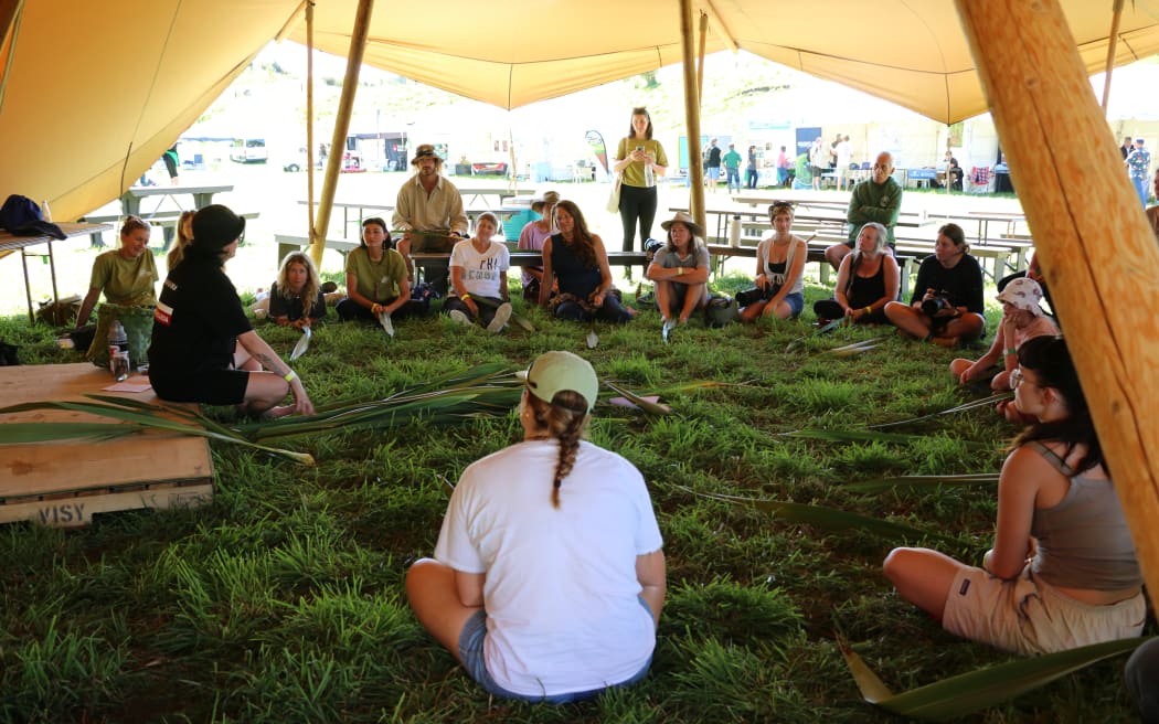 People sit in a circle, watching one woman on the left weaving strands of flax.
