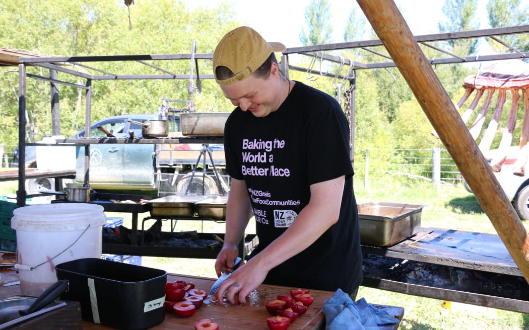Max Gordy cuts up fresh fruit. He is wearing a black shirt, a beige cap, and smiling.