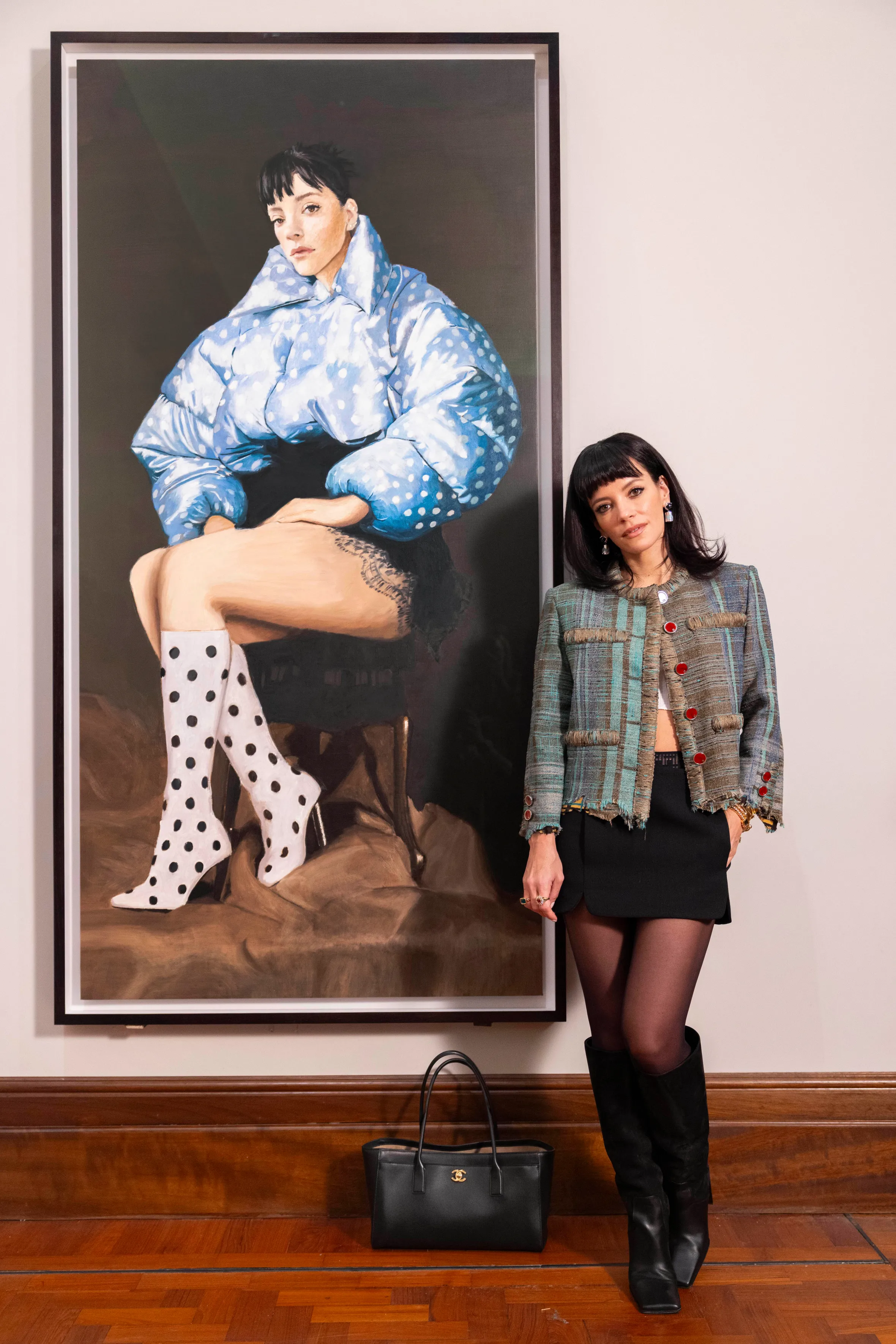 Lily Allen posing next to a painting of herself wearing a blue polka-dot puffer jacket and polka-dot boots.
