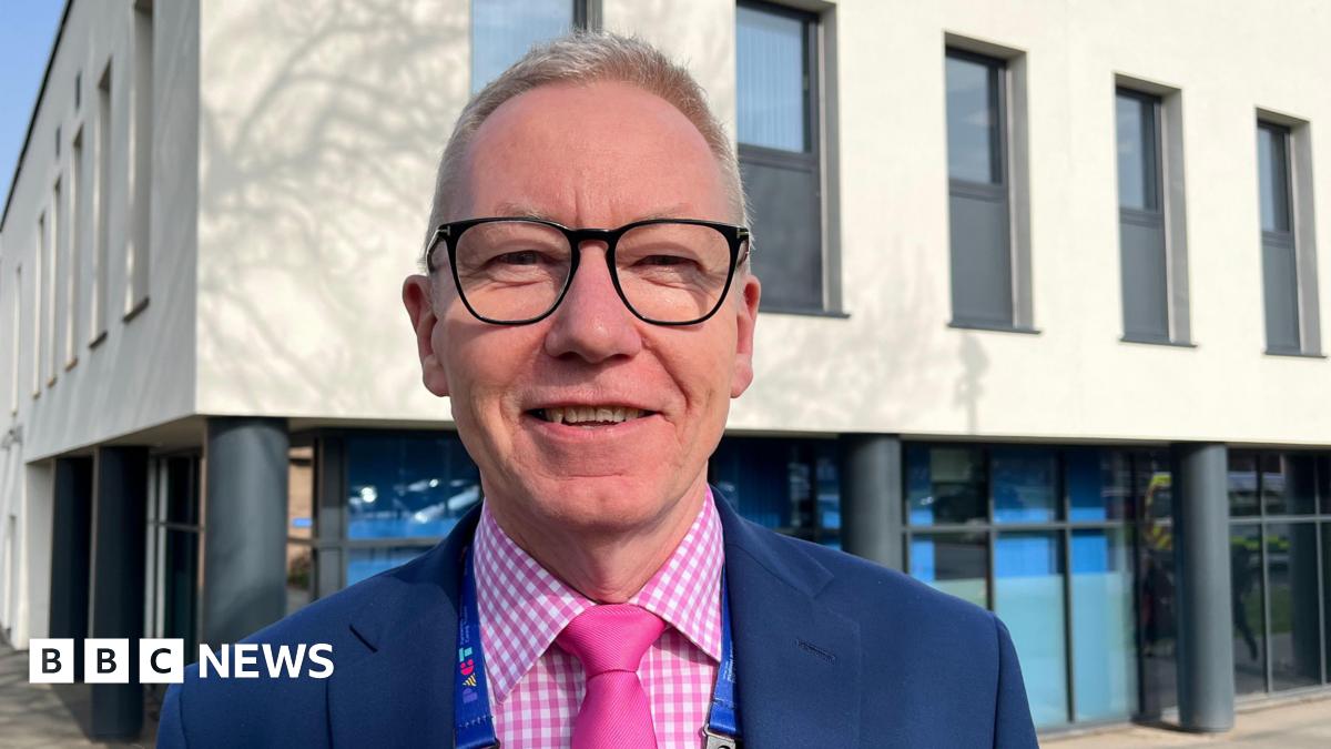 Andrew Morgan, who is looking into the camera and smiling. He is wearing a pink and white checked shirt, a pink tie, and a dark blue blazer. He has short, light blond hair and is wearing a pair of dark-framed glasses. Morgan is photographed outside a hospital building, which is a two-storey structure. The lower storey has dark grey pillars and floor-to-ceiling windows, whilst the upper floor is painted cream and has about eight, narrow, grey-framed windows visible.