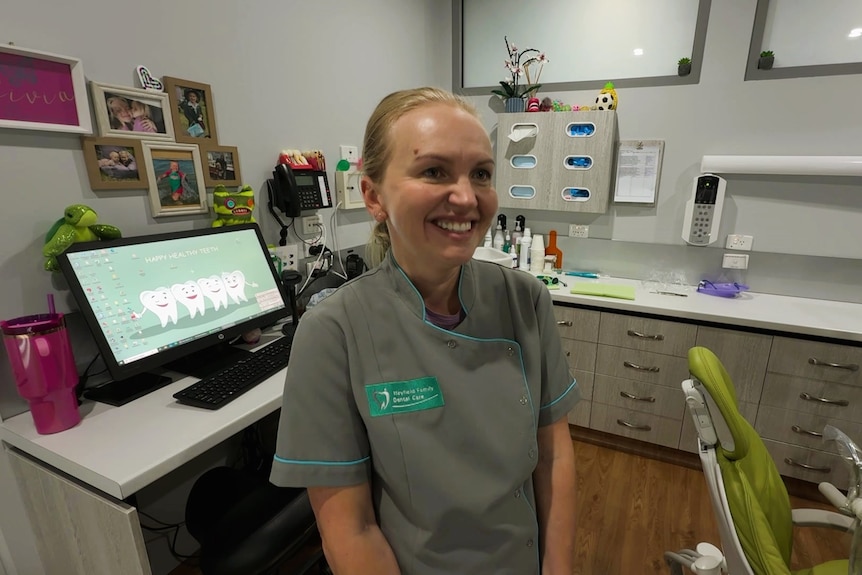 A photo of a blonde woman standing in a dentist theatre, with a dentist uniform and supplies in background.