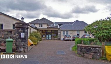 The hospital is made up of a number of low buildings with peaked, slate roofs. Three cars are parked near the glass entrance.