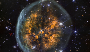 A shell of ghostly gas encapsulates a cloud of amber-colored gases that blow out in both directions from a central point. This makes the nebula look like a top-down view of two brain hemispheres inside a transparent skull. The scene is decorated with multicolor dots of light, representing distant galaxies and stars. The stars shown here have six points, characteristic of Webb images.