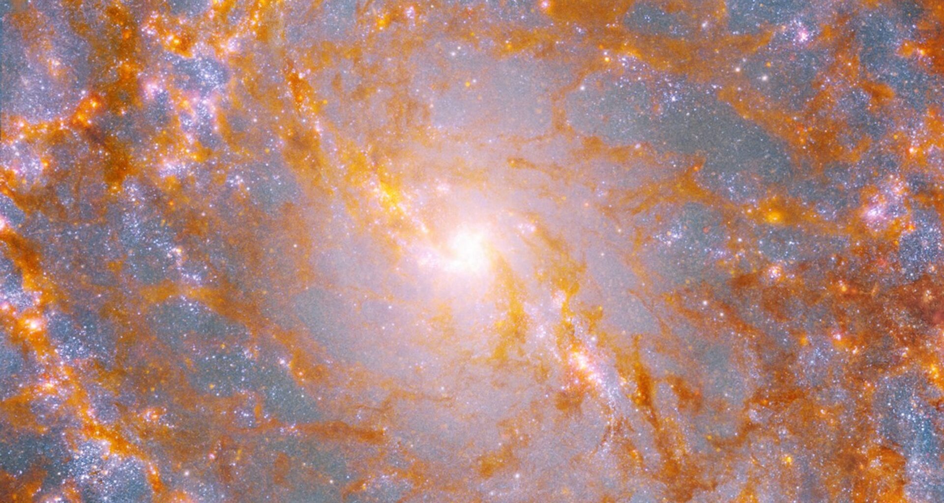 A close-up view shows a galaxy’s bright, yellow center, surrounded by patches of glowing orange-brown dust and pink stars.
