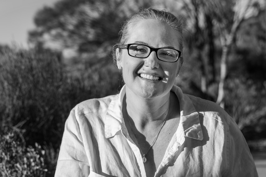 A black-and-white photo of a smiling woman wearing a white shirt with shrubland behind her.