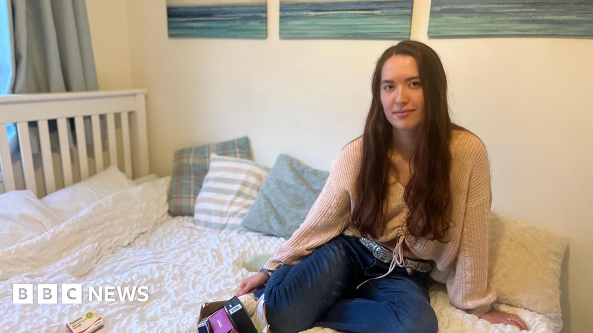 Lily, a young woman, is smiling. She has blonde hair and is wearing a grey woollen hat and scarf. In the background are skyscrapers and a river.
