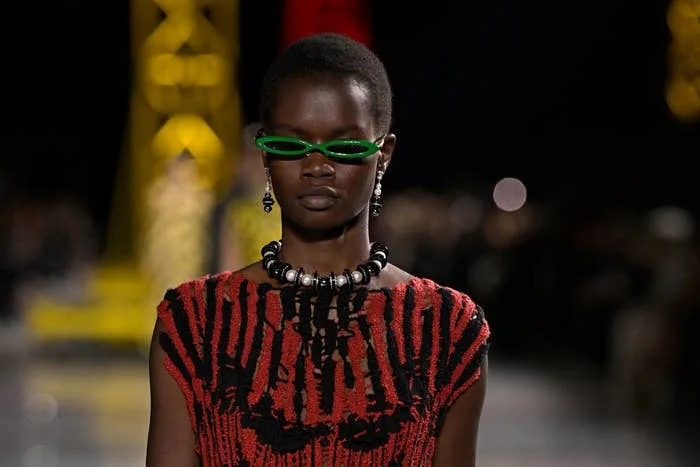 Model on runway wearing a striking textured dress with bold patterns, unique necklace, and futuristic sunglasses