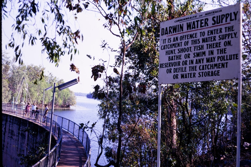A sign in the foreground gives swimmers instructions for dam use, behind it, is a bridge over water with people standing on it