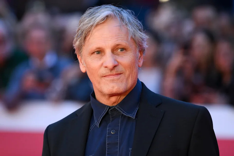 Viggo Mortensen in a dark jacket and shirt on a red carpet, with a crowd in the background