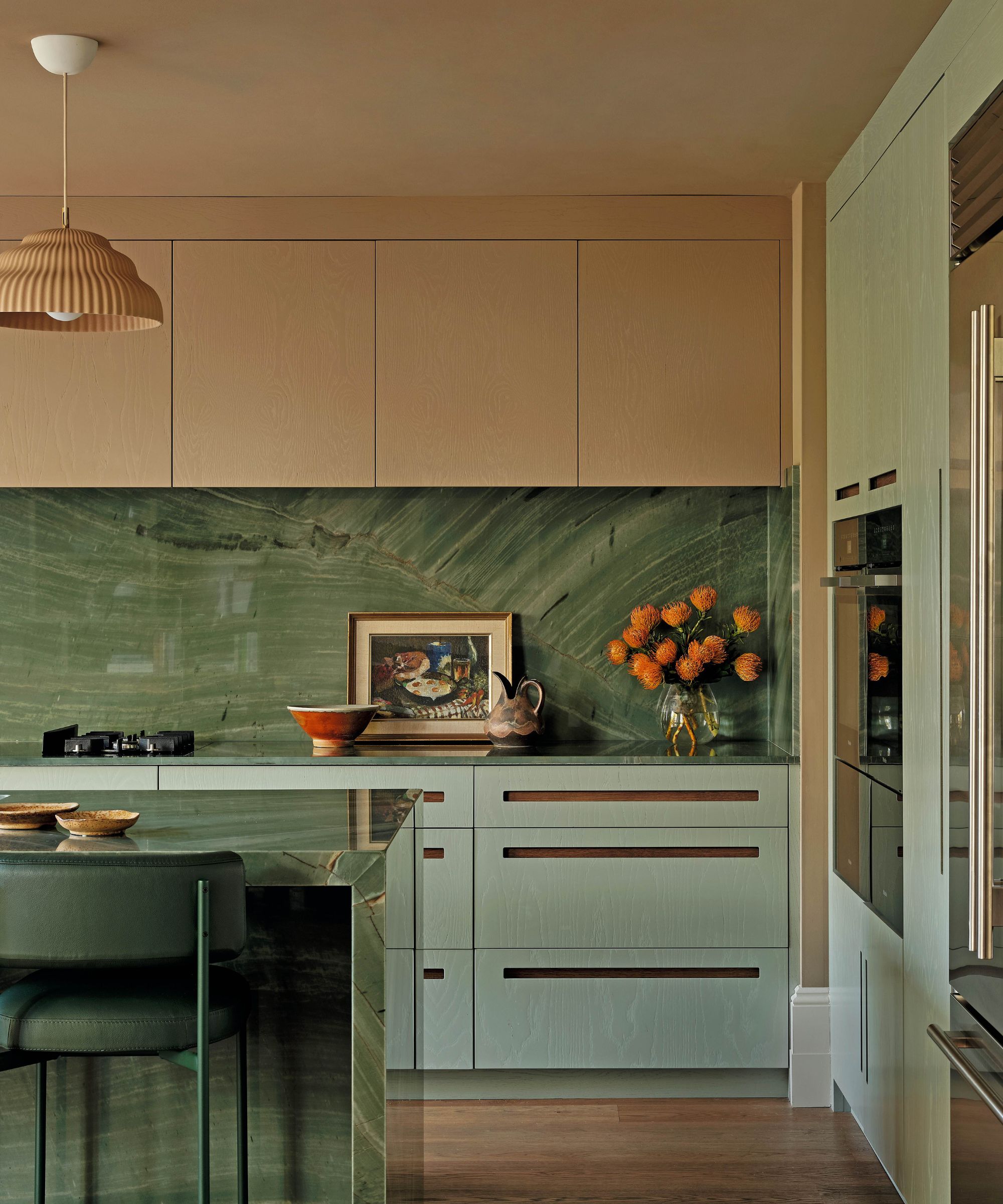 A contemporary kitchen showcasing sage green cabinetry with integrated wooden handles and a striking green marble backsplash and island. A fluted terracotta pendant light hangs above the workspace.