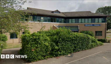 The outside of Elizabeth House, at Fulbourn Hospital. It is an angular brick building, with a line of windows on the top floor. In the foreground is a big green bush and in the distance, on the right, is a police car parked up.
