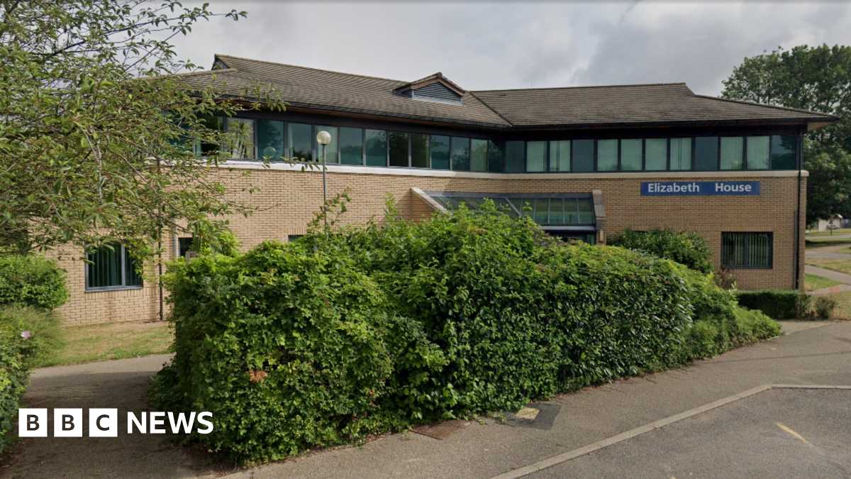 The outside of Elizabeth House, at Fulbourn Hospital. It is an angular brick building, with a line of windows on the top floor. In the foreground is a big green bush and in the distance, on the right, is a police car parked up.