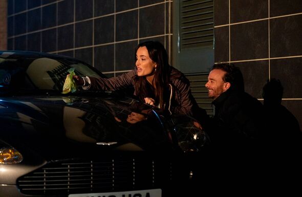 Two individuals are standing beside a sleek, dark vehicle in a dimly lit environment. The person on the left is positioned close