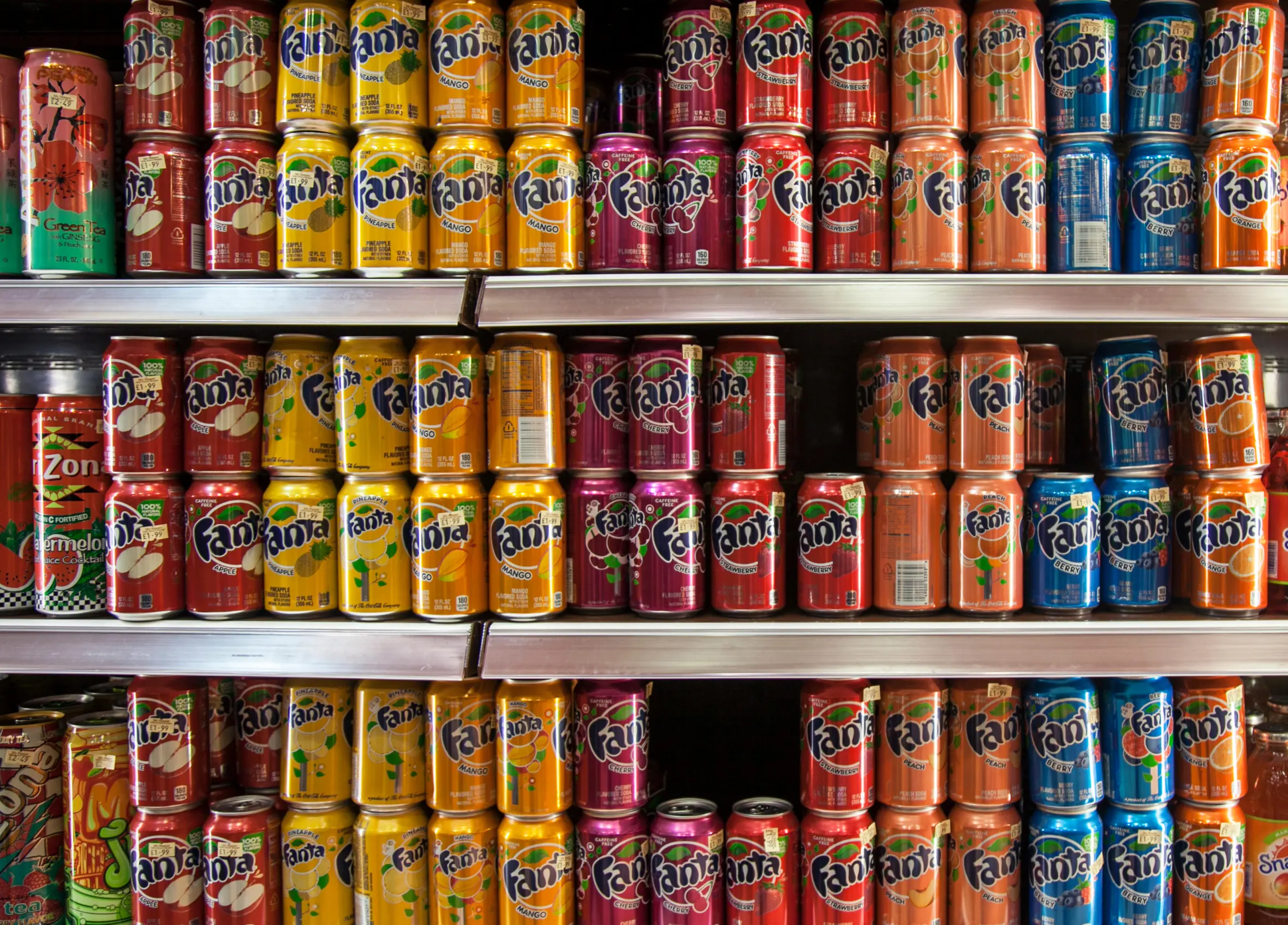 A display of Fanta soda cans in various fruit flavors, including pineapple, mango, cherry, strawberry, apple, peach, berry, and orange, stacked on shelves in a grocery store.