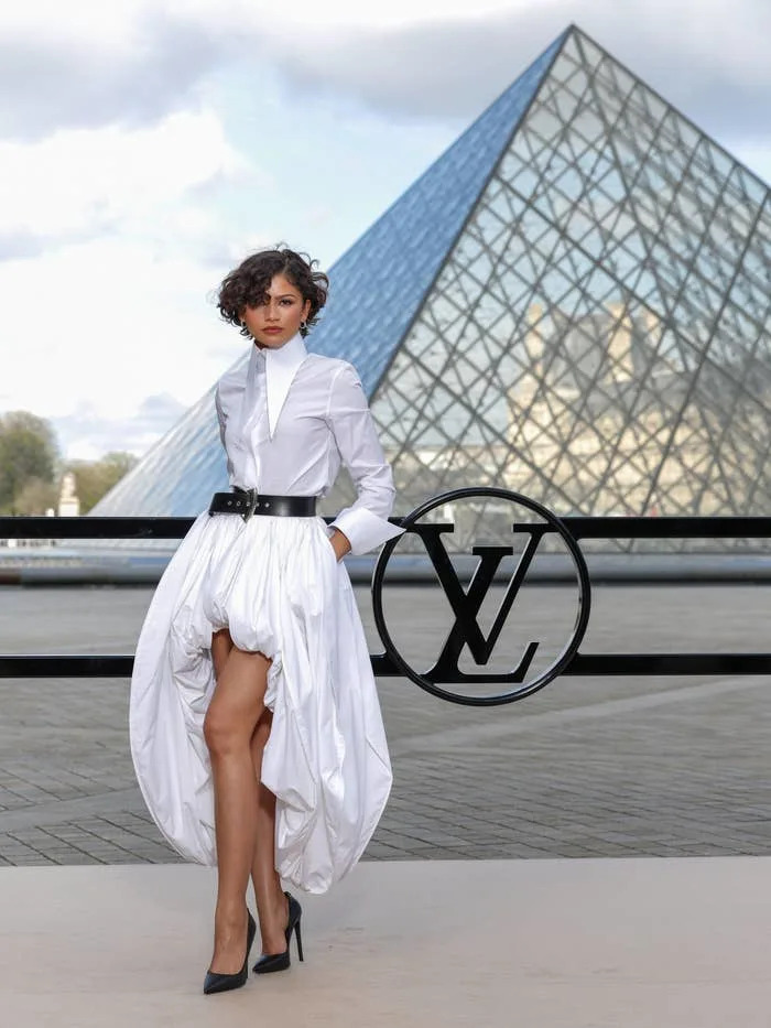Person poses elegantly in front of a pyramid structure, wearing a white high-low dress with a belt and black heels