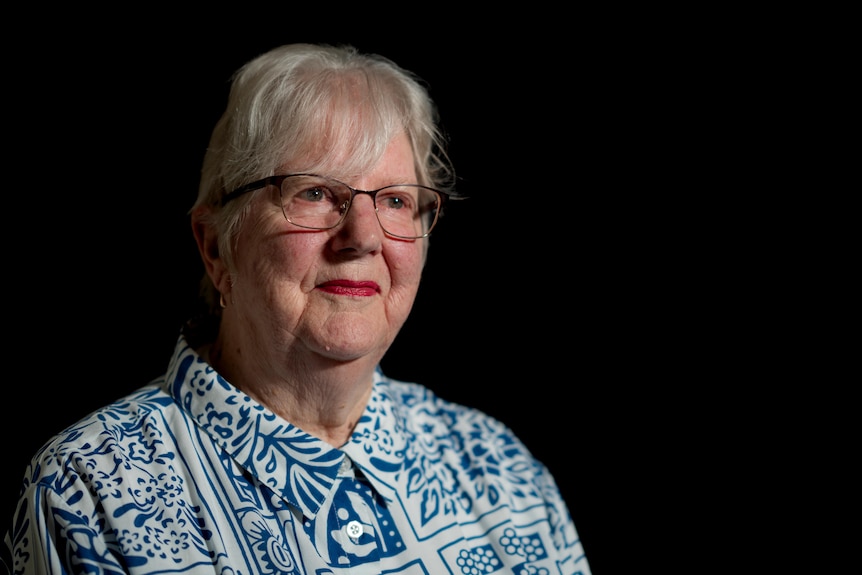 Suzenne, older woman with white hair, wearing a blue and white patterned top, glasses and red lipstick, on a black backdrop.