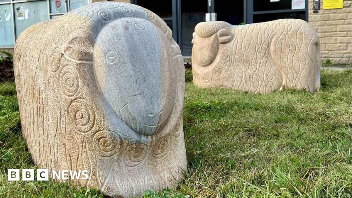 Two large carved stone sheep stand on a patch of grass in front of a brick building, each decorated with swirling patterns cut into their bodies. One sheep is darker and more detailed, while the other is lighter and simpler, with signs and windows from nearby offices visible behind them.