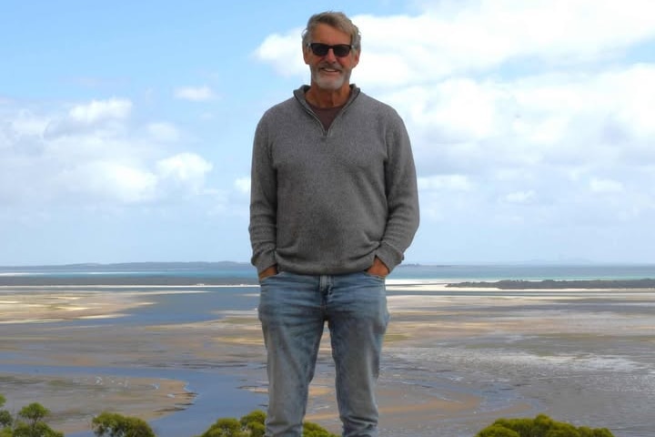 A man in a grey jumper and sunglasses stands at a coastal lookout overlooking a beach with the tide out